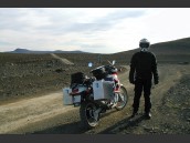 Person står bredvid parkerad MC (motorcykel) på grusväg i stenigt och kargt landskap på Island.