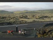 Motorcylist och två parkerade och semesterpackade motorcyklar vid väg på Island. Märkligt kalt och grönt landskap i kulissen.