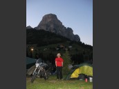 Person står mellan MC och tält på campingplats i Grödner Tal, Dolomiterna och Alperna på kvällen. Högt berg och blå himmel i bakgrunden.