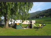 Två MC och tält under grönt träd på camping med stor grön gräsmatta i Mairatten, Alperna. Gröna skogsbevuxna berg och blå himmel i bakgrunden.