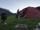 Kvällsvy från tält i marknivå med uppskuren korv, campingkök, ett annat tält, parkerad MC samt fjäll och blå himmel i kulissen.