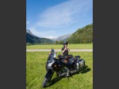Motorcyklist förbereder sig för körning vid svart MC, Suzuki V-Strom 650 på grön gräsmatta i Beiardalen, Nordland, Nordnorge, Norge. 
		Blå himmel med berg och snöklädda fjäll i bakgrunden.