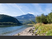 Strand och Beiarelva i Beiarn, Nordland, Nordnorge, Norge uppströms i vackert sommarväder. Berg och blå himmel med slöjmoln samt vita spår efter flygplan i bakgrunden.