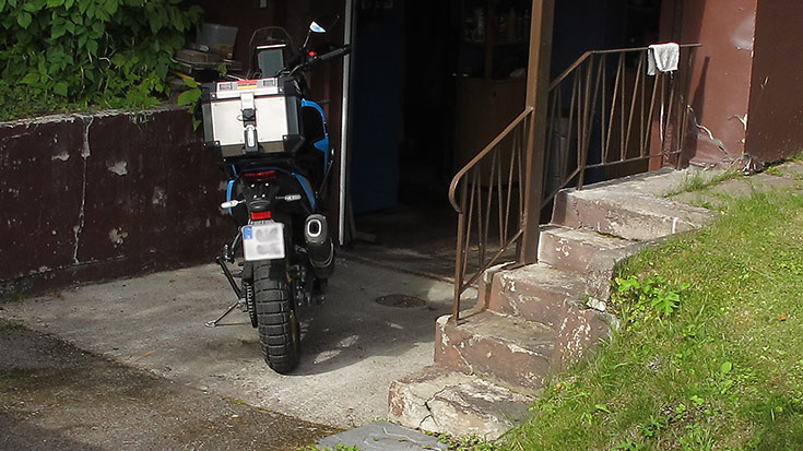 Blå MC, Yamaha Tenere 700, parkerad vid sprucken mur och husgrund vid en gargageinfart.